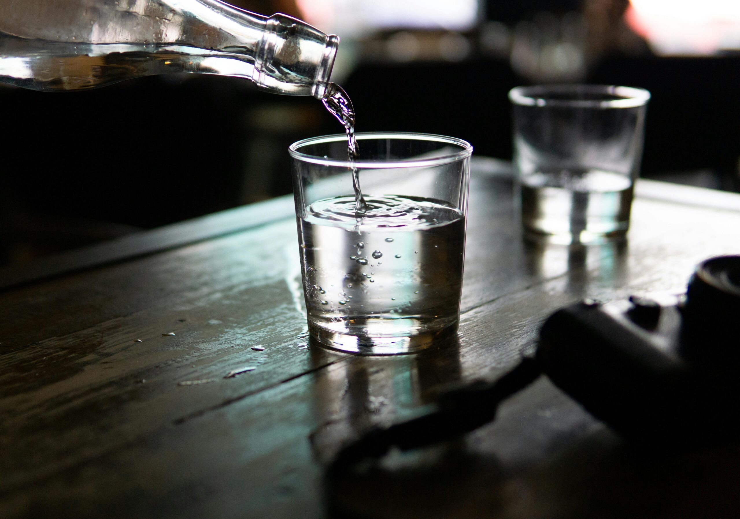 A clear glass of water being poured from a bottle, set on a rustic wooden table in a dimly lit indoor setting.
