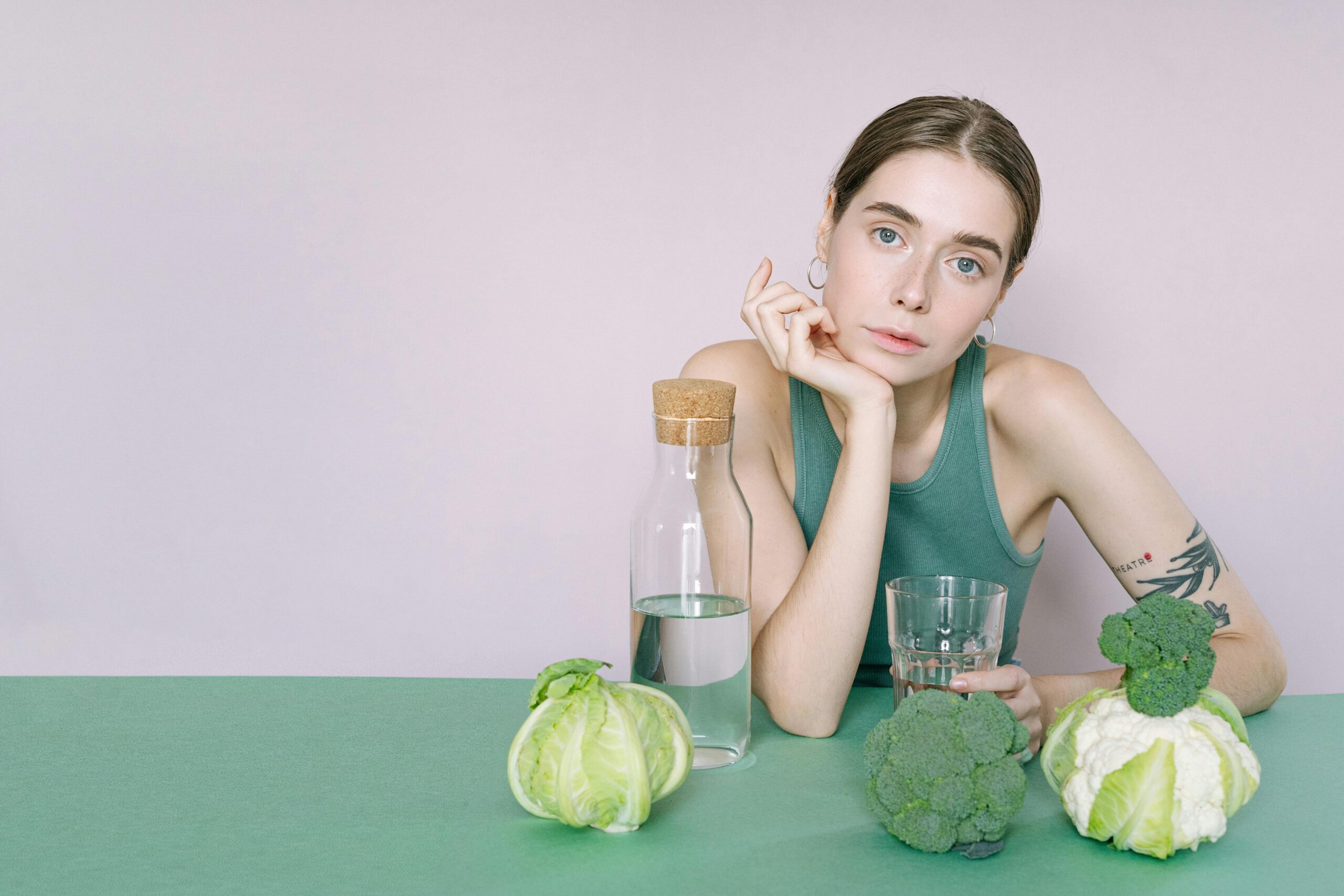 A young woman in a green top sitting with vegetables and a glass of water.