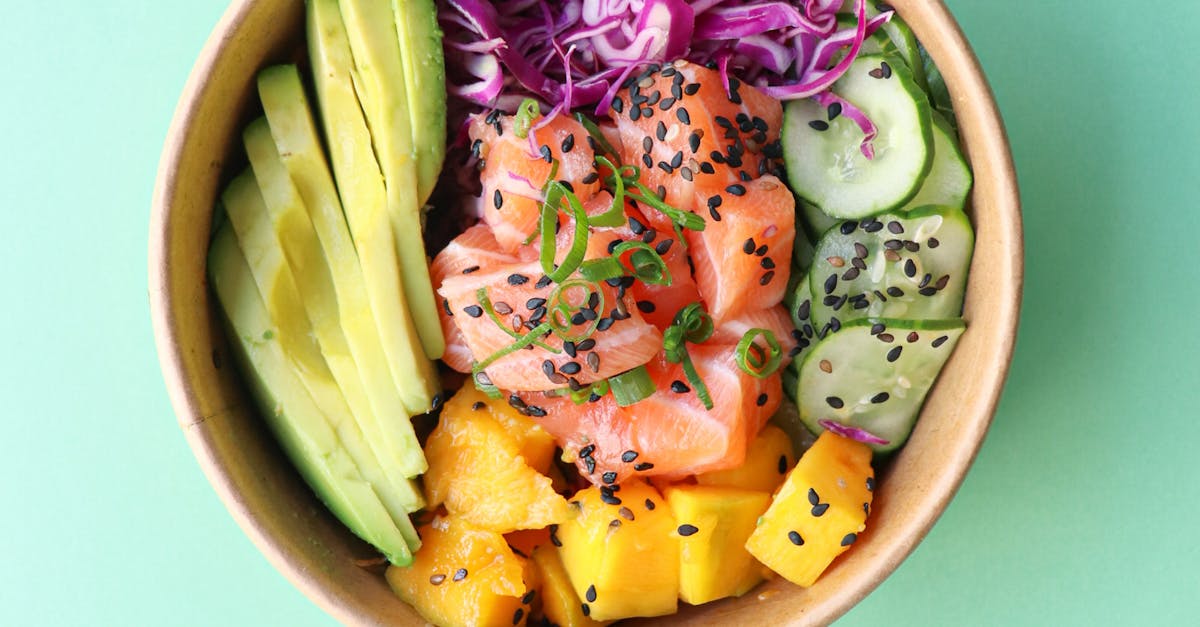 Top view of a vibrant poke bowl with salmon, avocado, mango, and vegetables on a mint background.